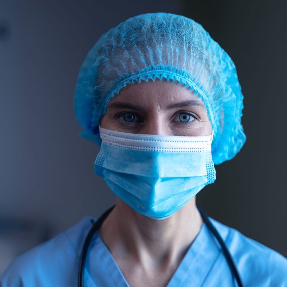 Portrait of caucasian female doctor in hospital wearing face mask, medical cap and scrubs. medical professional at work during coronavirus covid 19 pandemic.